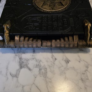 May include: A black decorative box with gold accents, featuring an ancient Egyptian theme. The box has a circular emblem and hieroglyphic-style carvings. The handle is a series of gold-colored rings. The box is resting on a white marble surface.
