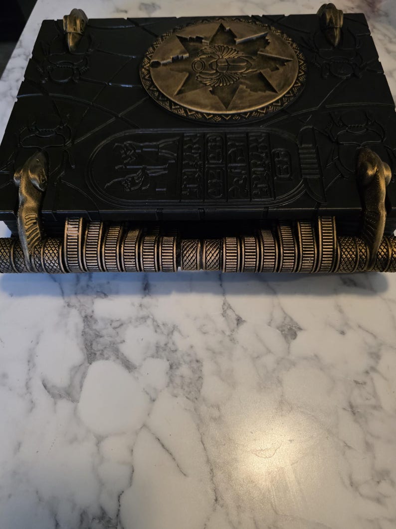 May include: A black decorative box with gold accents, featuring an ancient Egyptian theme. The box has a circular emblem and hieroglyphic-style carvings. The handle is a series of gold-colored rings. The box is resting on a white marble surface.
