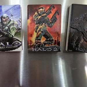 Könnte beinhalten: Drei rechteckige Halo-Spielelemente. Das erste zeigt einen Soldaten mit dem Text "HALO COMBAT EVOLVED". Das zweite zeigt einen Soldaten mit dem Text "HALO 2". Das dritte zeigt einen Soldaten mit dem Text "HALO FALL 2007".