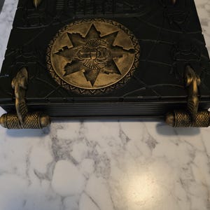 May include: A black decorative box with gold accents. The box features a central gold emblem with a star design and hieroglyphic-style detailing. Gold cobra-shaped handles and corner accents add to the ancient Egyptian theme.