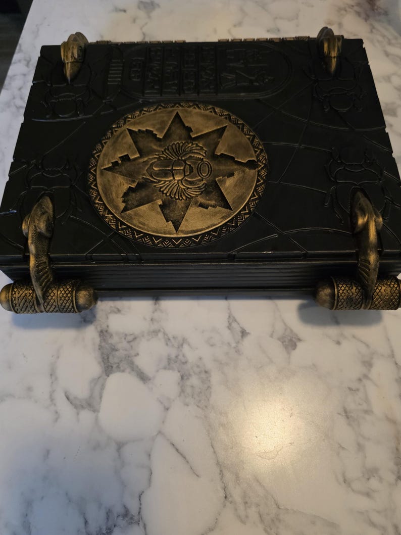 May include: A black decorative box with gold accents. The box features a central gold emblem with a star design and hieroglyphic-style detailing. Gold cobra-shaped handles and corner accents add to the ancient Egyptian theme.