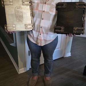 May include: Two large, ornate books with decorative metal clasps and trim. One book is light-colored with hieroglyphic carvings, the other is dark brown. The books are held by a person.