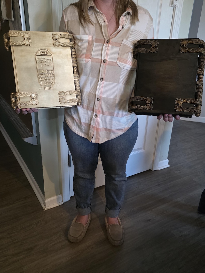 May include: Two large, ornate books with decorative metal clasps and trim. One book is light-colored with hieroglyphic carvings, the other is dark brown. The books are held by a person.
