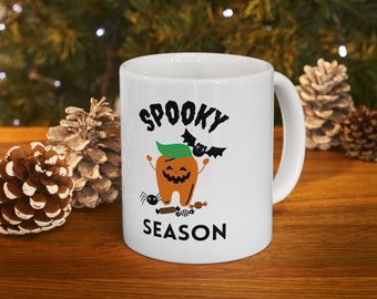 May include: White ceramic mug with a black and orange Halloween themed design. The mug features a cartoon tooth with a pumpkin face, a bat, and the text "Spooky Season".
