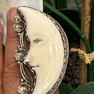 May include: A crescent moon-shaped pendant with a carved face, set in a silver-toned frame. The moon has a cream-colored surface and is adorned with floral accents. The pendant is held by a hand.