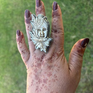 May include: A silver-toned ring featuring a carved ivory-colored Buddha face, framed by a lotus flower and leaves. The ring is set against a background of green grass. The ring is a unique piece of jewelry.