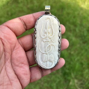May include: An oval pendant featuring a cream-colored carved relief of a trident and drum symbol set within a decorative silver-toned metal frame with a rope-like border and a top bail for hanging, showcasing intricate craftsmanship and detail.