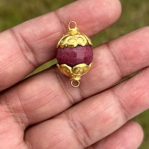 May include: A gold-colored pendant with a faceted, oval-shaped, deep red gemstone. The gold setting features ornate, petal-like designs at the top and bottom, with small loops for hanging. The pendant is held in a hand against a blurred green background.