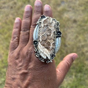 May include: An ornate, oversized ring with a detailed, textured design. The ring features a central, cream-colored oval with a raised pattern, framed by a silver-toned metal setting. Decorative elements include feathers and small black flower accents.
