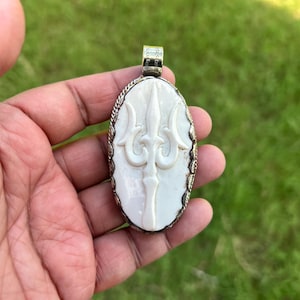 May include: An oval pendant featuring a white carved trident symbol set against a smooth background. The pendant is encased in a decorative metallic frame with a textured edge and includes a small silver-toned bail at the top for attachment.