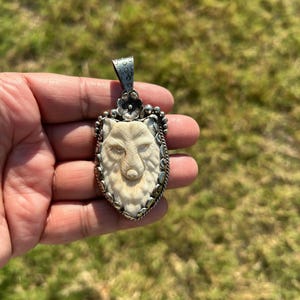 May include: Oval pendant featuring a carved wolf head in cream color, set within a silver-toned frame. The pendant has a detailed wolf face with a floral accent at the top and a textured bail for a chain.