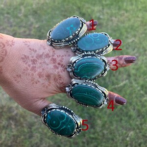 May include: Five silver rings, each with a large, oval, green malachite stone, are shown on a hand. The rings have decorative silver settings with leaf and floral details. The malachite stones display unique swirling patterns.