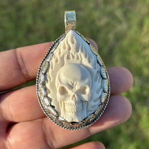 May include: A pendant featuring a carved skull design within a teardrop shape. The skull is cream-colored, surrounded by a flame-like pattern, and framed with a silver-colored metal border. The pendant has a gold-colored bail.