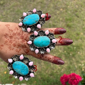May include: Three silver-toned rings with turquoise oval stones and pink and silver floral accents. The rings are displayed on a hand with henna designs and red nail polish, set against a blurred green background.