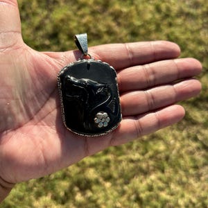 Pode incluir: Um pingente retangular preto com borda e argola prateadas. O pingente apresenta uma imagem esculpida da cabeça de um lobo e uma pequena flor. O pingente é segurado em uma mão contra um fundo verde desfocado.