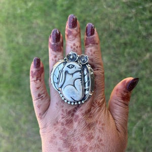 Op de afbeelding: Zilveren ring met een konijnontwerp en bloemendetails. De ring heeft een ovale voorkant met een verhoogd konijnmotief en een decoratieve rand. De hand draagt henna-patronen en donkerrode nagellak.