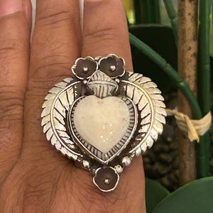 May include: A large silver ring featuring a heart-shaped, off-white stone at its center. The ring is surrounded by a detailed silver design, including leaves and small flower accents. The ring is on a hand.