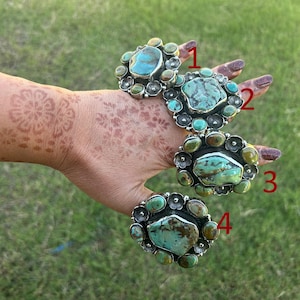 May include: Four silver rings with large turquoise stones are displayed. Each ring features a unique turquoise stone surrounded by smaller, decorative elements. The rings are set against a backdrop of green grass.