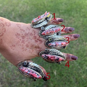May include: Five silver rings with oval, pink and red stone settings are displayed on a hand. The rings have a leaf-like design around the stone. The background is blurred green grass.
