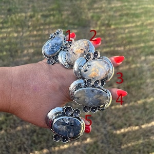 May include: Five silver rings labelled one to five, each featuring a unique oval-shaped dendritic agate stone. The metal settings are decorated with leaf and floral motifs. The stones display various patterns of dark inclusions against translucent backgrounds.