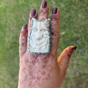 May include: A silver ring with a carved wolf head design. The wolf has detailed facial features and is set against a rectangular background. The ring is worn on a hand with dark red nail varnish.