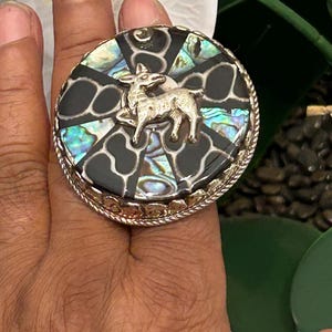 May include: A large, round ring with a silver-colored metal band. The ring's face features a black and iridescent abalone inlay with a silver-colored deer figure in the center. The ring has a textured, rope-like border.