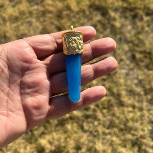 Könnte beinhalten: Ein blauer Kristallanhänger mit einer goldfarbenen, verzierten Kappe. Der Anhänger wird in einer Hand vor einem verschwommenen Hintergrund aus Gras gehalten. Der Kristall hat eine leuchtend blaue Farbe und ein spitzes Ende.