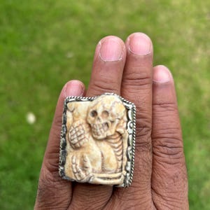 May include: A rectangular ring with a detailed skull carving. The ring has a silver-colored border and a cream-colored skull design. The ring is held up by a hand against a green background.