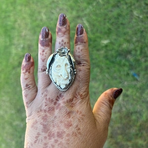 May include: A silver ring with a carved, off-white wolf face in an oval frame. The ring has a detailed, textured design and is worn on a hand with henna patterns and burgundy nail polish. The background is blurred green grass.