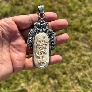 May include: A silver pendant with an ornate design. The pendant features a central ivory-colored oval with carved symbols and is framed by a detailed silver border. The pendant is held up by a silver loop.