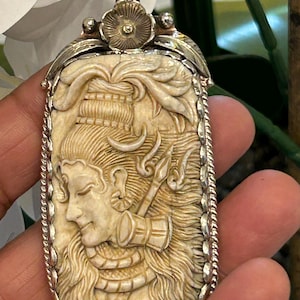 May include: A cream-coloured rectangular pendant with a detailed carving of a deity, set within a silver-coloured frame and bail. The carving depicts the deity's face, hair, and accessories with intricate detail. The pendant features a floral design at the top.