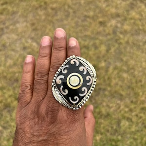 Könnte beinhalten: Ein großer, verzierter Ring mit einem schwarzen, quadratischen Mittelstück mit weißen und cremefarbenen Wirbelmustern. Der Ring hat einen silberfarbenen, strukturierten Rand mit blattartigen Details. Der Ring befindet sich an einer Hand.