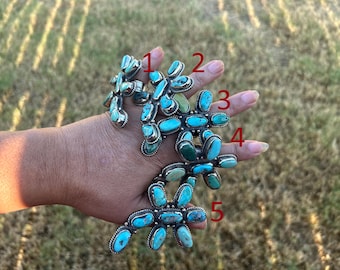 May include: Five silver rings featuring clusters of oval-shaped turquoise stones in various shades of blue and green. Each ring is numbered one through five in red text, showcasing intricate metalwork settings that hold multiple stones in place.