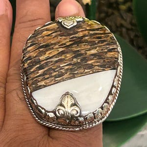 May include: Oval-shaped ring with a silver band. The ring features a two-tone design: a section of brown and tan wood-like material and a section of white stone. Decorative silver accents are present on the ring's surface.