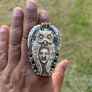 Könnte beinhalten: Ein großer, verzierter Ring mit einem geschnitzten Eulen-Design und einem Frauengesicht in der Mitte. Der Ring hat ein silberfarbenes Band und eine detaillierte, cremefarbene Eule und ein Gesicht. Der Ring wird an einer Hand getragen.