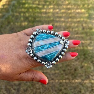 May include: A large statement ring featuring a triangular face with alternating diagonal stripes of crushed turquoise-colored stone and iridescent shell inlay. The setting is polished silver with a beaded border and small flower accents.