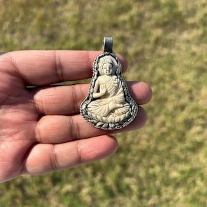 May include: A pendant featuring a carved ivory-colored Buddha figure seated in a meditative pose. The Buddha is framed by a decorative silver-toned border with a loop for a chain. The pendant is held in a hand against a blurred green background.