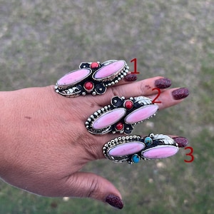May include: Three silver rings with oval pink stones, red and turquoise accents. The rings have intricate metalwork and are displayed on a hand with dark red nail polish. The rings are set against a blurred green background.