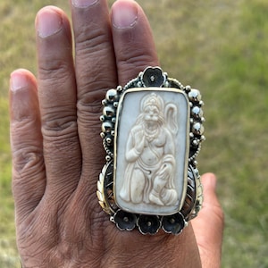 May include: A large, ornate ring with a rectangular ivory-colored centerpiece featuring a carved figure. The ring has a silver-colored setting with floral and beaded accents. The ring is on a hand.