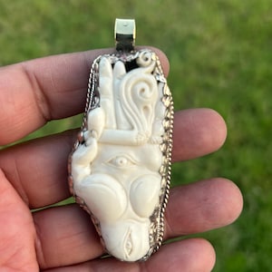 Può includere: Ciondolo color avorio con un'intagliatura dettagliata di una mano con un occhio e altri simboli. Il ciondolo è incastonato in una cornice di metallo color argento con un'ansa dorata. Lo sfondo è verde sfocato.