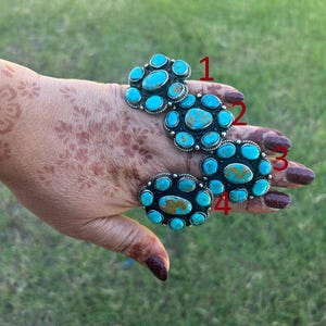 May include: Four silver-toned rings with turquoise stones. Each ring features a cluster of bright blue turquoise stones, with some rings having a central oval stone. The rings are displayed on a hand with henna designs.