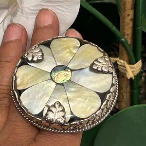 May include: A round, silver-toned jewelry box with a floral design. The lid features mother-of-pearl petals and a central abalone accent. Decorative leaf accents are placed around the edge. The box is held in a hand.