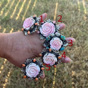 May include: Five silver rings labeled 1 through 5, each featuring a carved light pink rose centerpiece. The rings are accented with small orange or blue stones set in decorative silver metalwork, arranged in a vertical sequence on a hand.