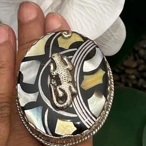 May include: A round, silver-toned pendant with a detailed lizard design. The pendant's face features a mosaic of black, white, and yellow iridescent shell pieces. The border has a twisted rope design.