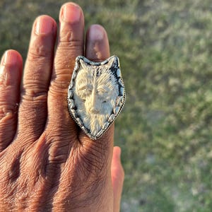 May include: A silver ring with a carved wolf head design. The wolf's face is detailed with fur and facial features, set against a textured background. The ring has a decorative border.