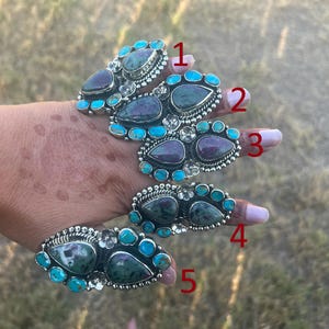 May include: Five silver rings labeled 1 through 5, each featuring teardrop-shaped dark purple and green stones surrounded by small round turquoise cabochons and silver floral accents. The rings are arranged on fingers, showcasing intricate metalwork.