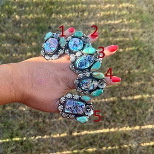 May include: Five silver rings labeled one through five, each featuring iridescent abalone shell inlays and small turquoise stones. The metal settings have decorative floral accents and scalloped edges, showcasing a detailed, ornate craftsmanship.