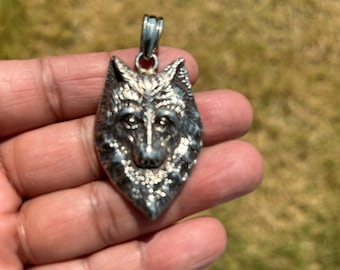 May include: A silver wolf pendant with detailed features. The pendant depicts a wolf's head with a textured surface and a loop for a chain. The pendant is held in a hand against a blurred green background.