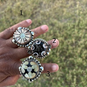 May include: Three silver rings with distinctive designs. Ring 1 has a cream-coloured centre with a sunburst pattern. Ring 2 features a black centre with a white star design. Ring 3 has a black centre with a white floral design. All rings have a beaded border.