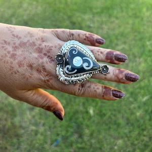May include: A silver ring with a large, triangular black stone with white swirls. The ring has a decorative silver border and small flower accents. The ring is on a hand with henna designs and dark red nail polish, set against a green background.